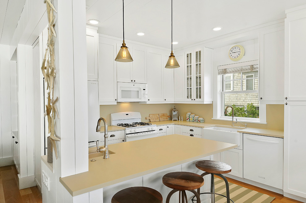 Photo of a small beach style l-shaped eat-in kitchen in Other with a farmhouse sink, shaker cabinets, white cabinets, solid surface benchtops, beige splashback, white appliances, light hardwood floors and a peninsula.