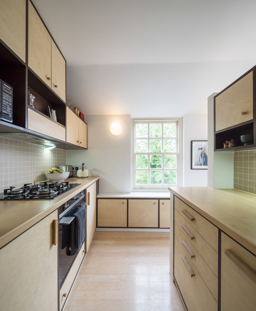 This is an example of a contemporary galley kitchen in London with flat-panel cabinets, light wood cabinets, wood benchtops, beige splashback, ceramic splashback, black appliances, light hardwood floors and no island.