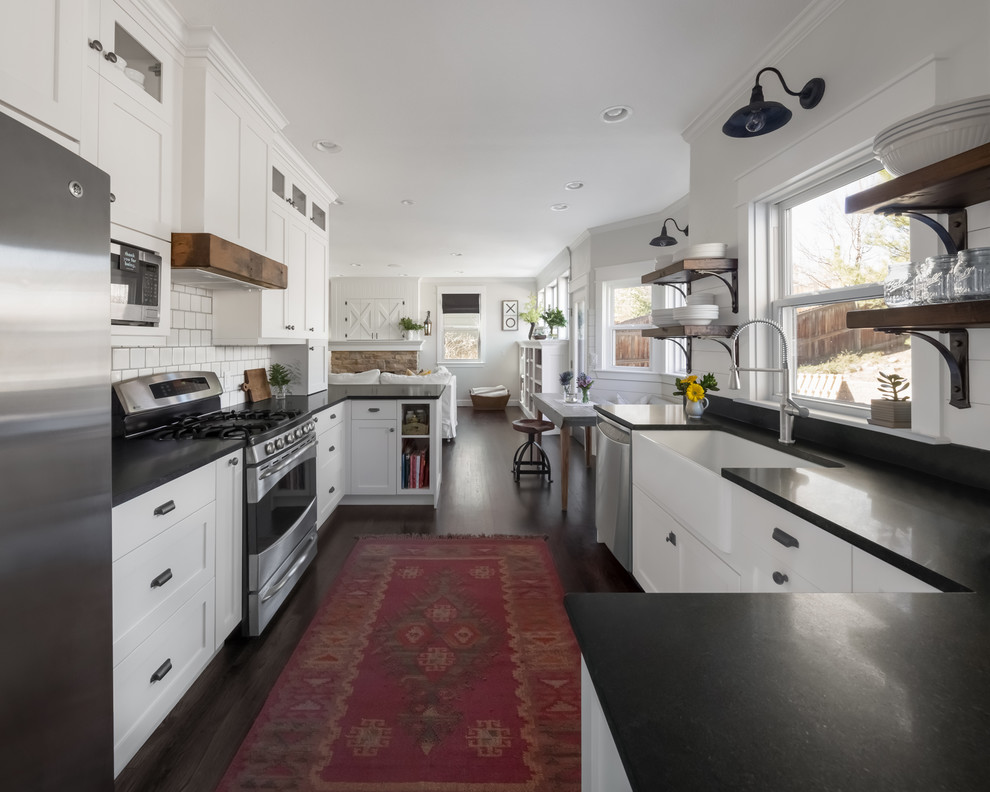 Design ideas for a mid-sized country galley eat-in kitchen in Denver with a farmhouse sink, shaker cabinets, white cabinets, granite benchtops, white splashback, ceramic splashback, stainless steel appliances, dark hardwood floors, a peninsula, brown floor and black benchtop.