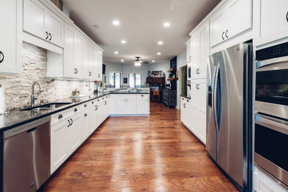Example of a large transitional galley light wood floor and brown floor open concept kitchen design with an undermount sink, shaker cabinets, white cabinets, quartz countertops, white backsplash, stone tile backsplash, stainless steel appliances and a peninsula