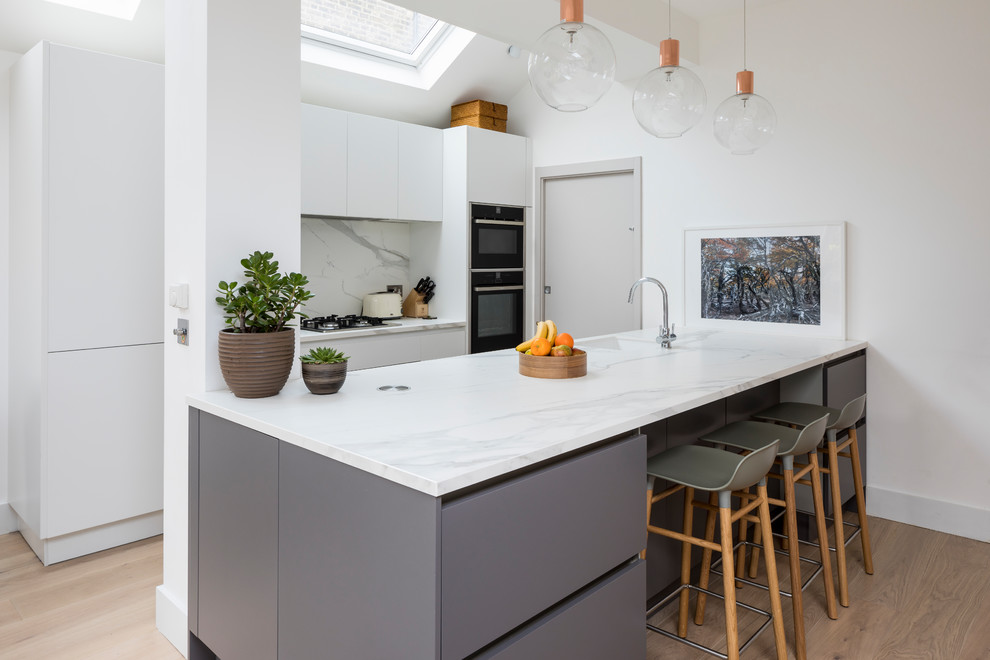 This is an example of a contemporary galley kitchen in London with an undermount sink, flat-panel cabinets, white cabinets, grey splashback, stone slab splashback, black appliances, light hardwood floors, a peninsula and beige floor.