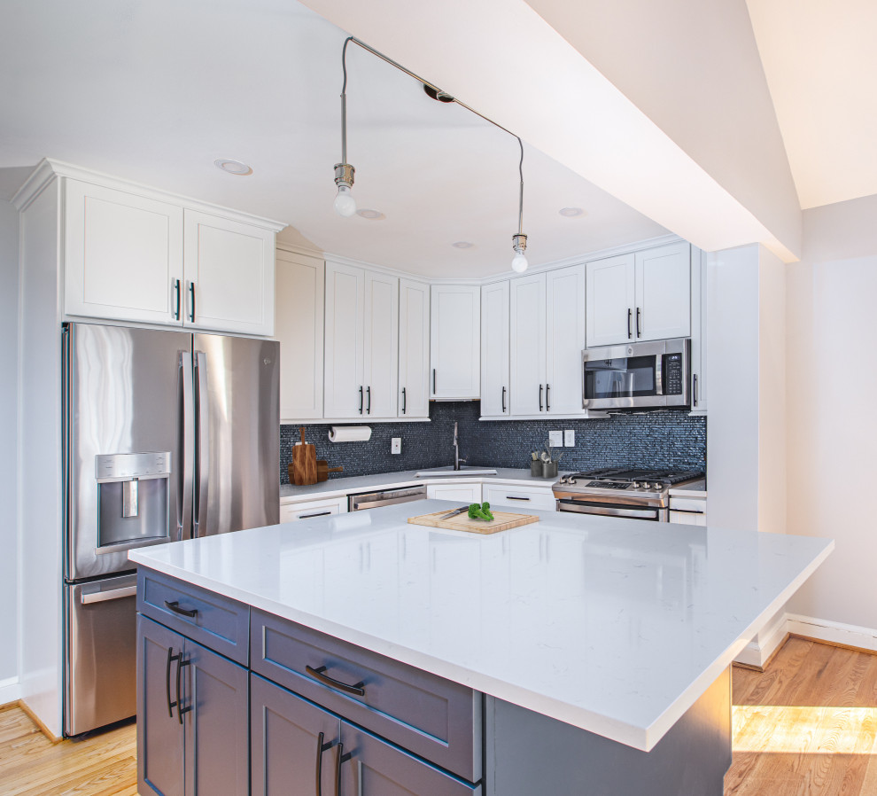 Small contemporary l-shaped kitchen/diner in DC Metro with a submerged sink, shaker cabinets, white cabinets, engineered stone countertops, blue splashback, mosaic tiled splashback, stainless steel appliances, light hardwood flooring, an island, brown floors and white worktops.