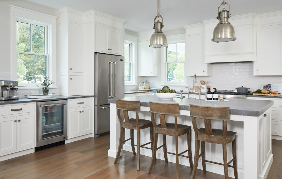 Mid-sized country l-shaped open plan kitchen in Grand Rapids with a farmhouse sink, recessed-panel cabinets, white cabinets, concrete benchtops, white splashback, subway tile splashback, stainless steel appliances, medium hardwood floors, with island and grey benchtop.