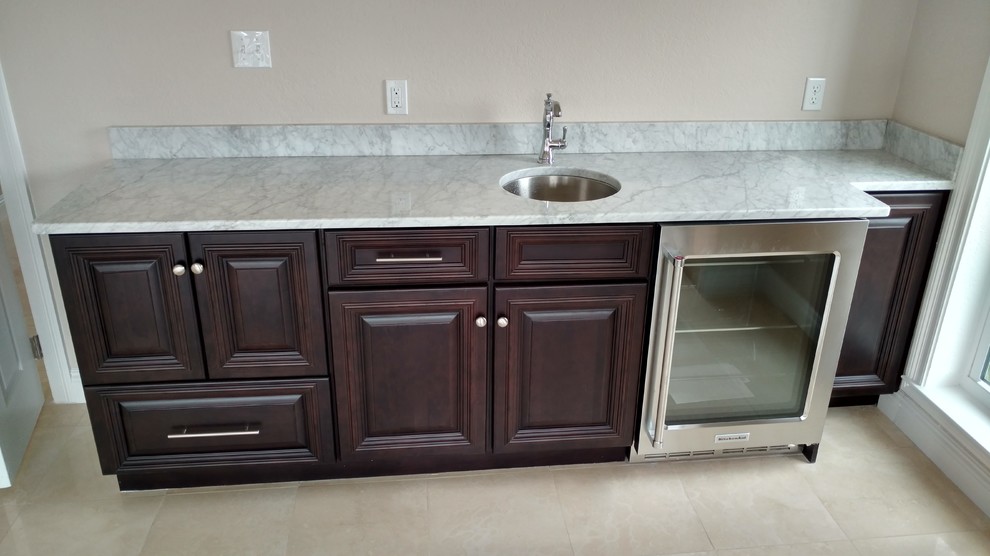 Inspiration for a small beach style galley wet bar in Miami with an undermount sink, raised-panel cabinets, dark wood cabinets, marble benchtops, white splashback and travertine floors.