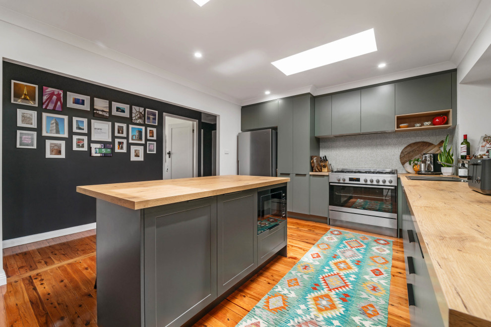 Photo of a mid-sized contemporary l-shaped kitchen pantry in Sydney with a drop-in sink, flat-panel cabinets, green cabinets, wood benchtops, grey splashback, cement tile splashback, stainless steel appliances, light hardwood floors, with island and brown benchtop.