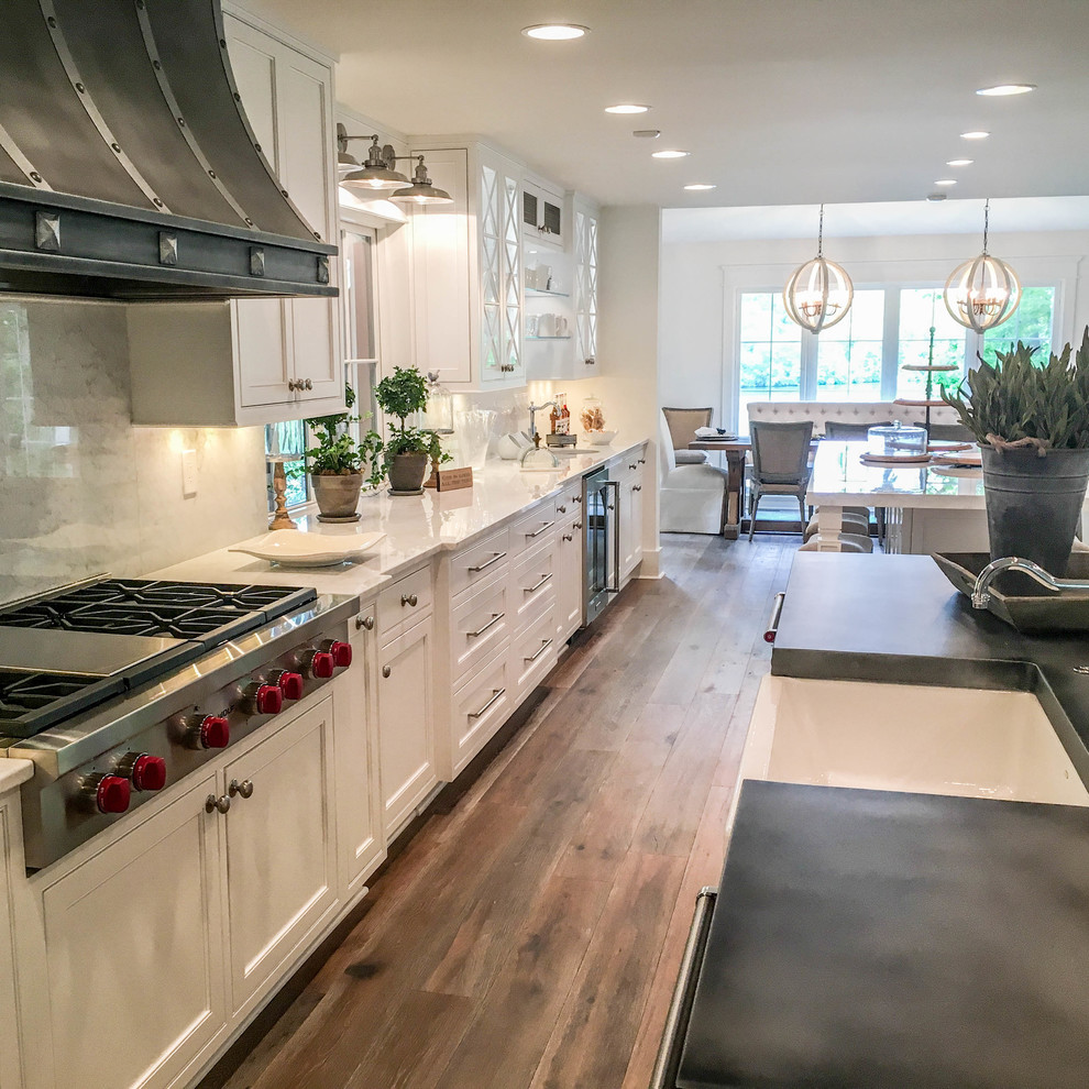 Photo of an expansive transitional l-shaped kitchen in Other with a farmhouse sink, beaded inset cabinets, white cabinets, zinc benchtops, white splashback, marble splashback, stainless steel appliances, light hardwood floors, multiple islands and brown floor.