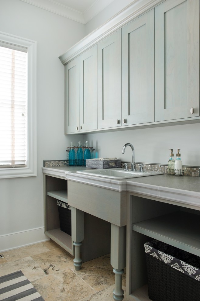 Photo of a mid-sized transitional galley utility room in Kansas City with a drop-in sink, shaker cabinets, grey cabinets, tile benchtops, grey walls, travertine floors and a side-by-side washer and dryer.