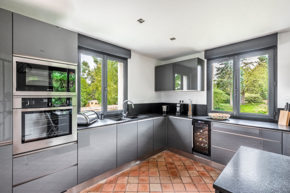 Photo of a contemporary l-shaped kitchen in Paris with a double-bowl sink, flat-panel cabinets, grey cabinets, black splashback, stainless steel appliances, terra-cotta floors, with island, orange floor and black benchtop.