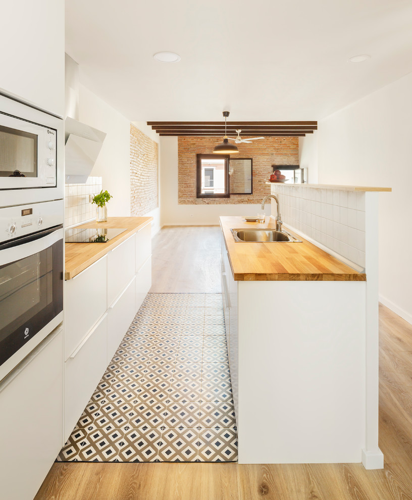 Photo of a mid-sized mediterranean galley open plan kitchen in Barcelona with a single-bowl sink, flat-panel cabinets, white cabinets, wood benchtops, white splashback, ceramic splashback, stainless steel appliances, ceramic floors and multi-coloured floor.
