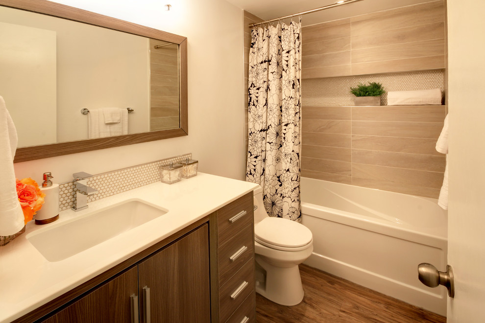 Photo of a small modern bathroom in Calgary with flat-panel cabinets, medium wood cabinets, an alcove tub, a shower/bathtub combo, a one-piece toilet, beige tile, ceramic tile, white walls, vinyl floors, an undermount sink, engineered quartz benchtops, brown floor and a shower curtain.