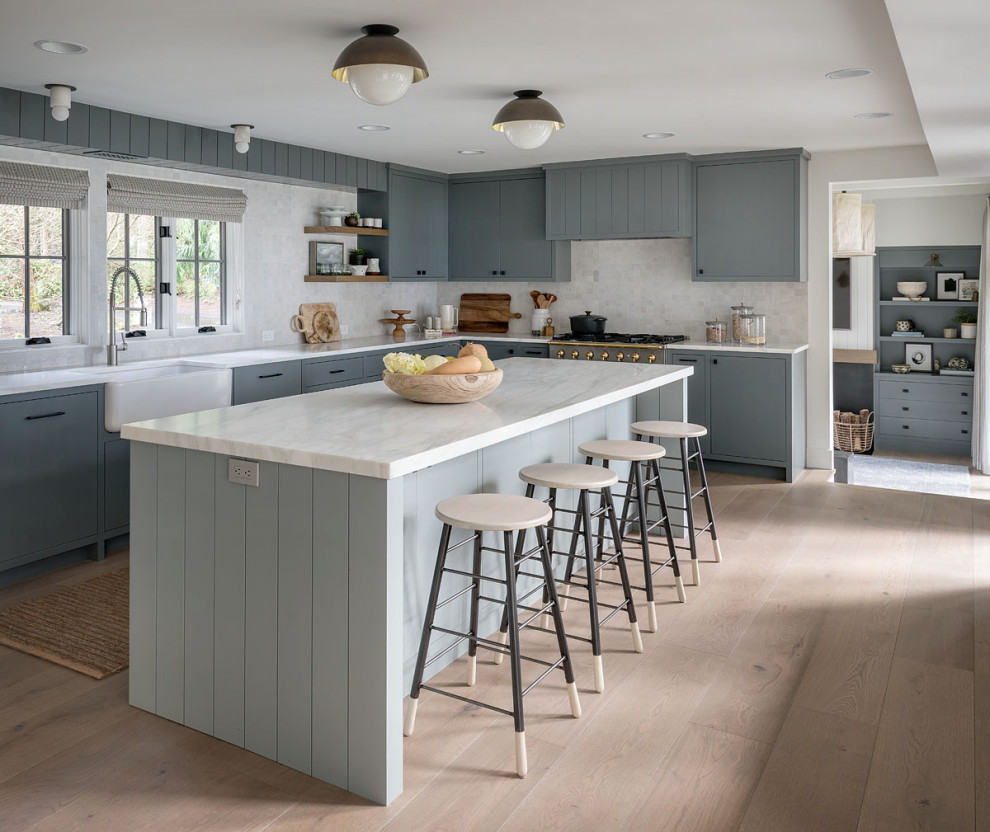 This is an example of a country l-shaped kitchen in Seattle with a farmhouse sink, flat-panel cabinets, white splashback, stainless steel appliances, light hardwood floors, with island, beige floor, white benchtop and blue cabinets.
