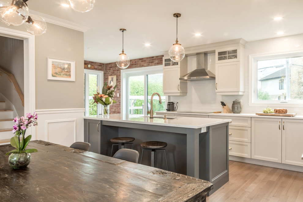 Eat-in kitchen - mid-sized transitional l-shaped medium tone wood floor and brown floor eat-in kitchen idea in Montreal with an undermount sink, recessed-panel cabinets, white cabinets, quartz countertops, white backsplash, ceramic backsplash, paneled appliances and an island