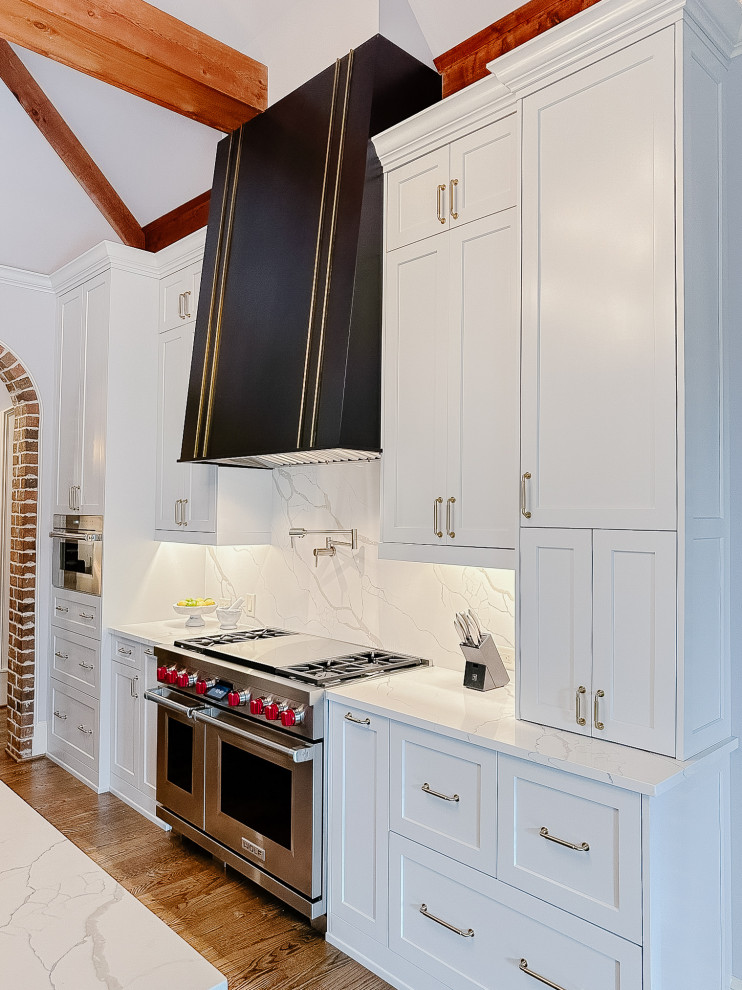 Inspiration for an expansive transitional l-shaped open plan kitchen in Atlanta with a single-bowl sink, shaker cabinets, white cabinets, quartz benchtops, white splashback, engineered quartz splashback, stainless steel appliances, medium hardwood floors, with island, brown floor, white benchtop and exposed beam.