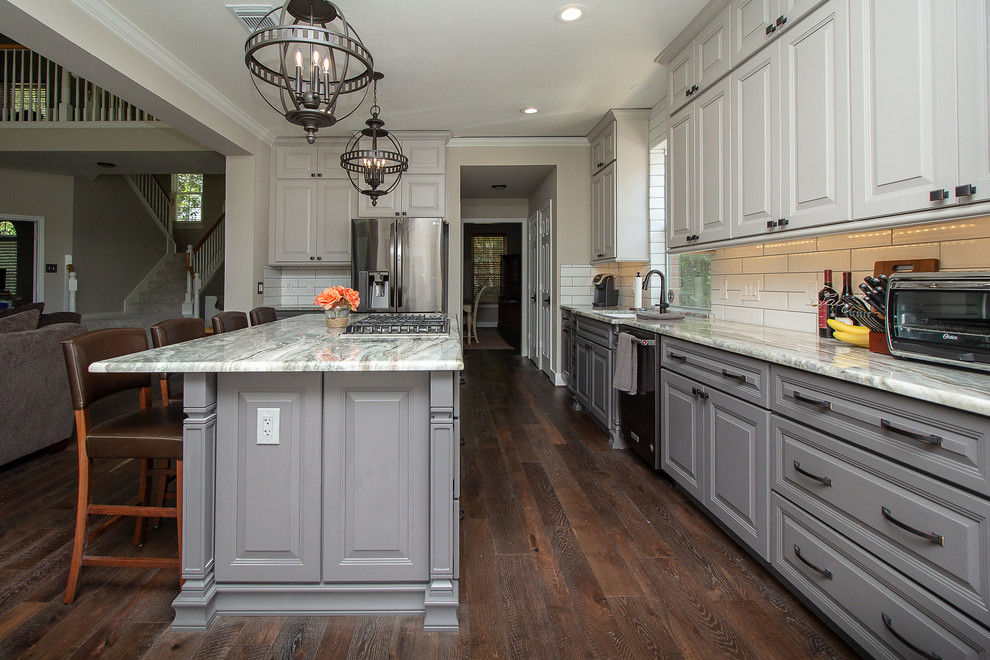 Large traditional l-shaped open plan kitchen in Houston with an undermount sink, raised-panel cabinets, grey cabinets, granite benchtops, white splashback, subway tile splashback, stainless steel appliances, medium hardwood floors, with island, brown floor and grey benchtop.