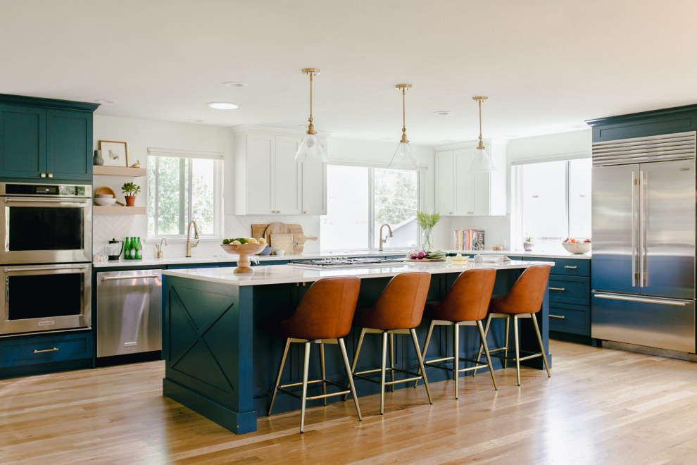 This is an example of a large transitional u-shaped open plan kitchen in Denver with an undermount sink, shaker cabinets, blue cabinets, white splashback, stainless steel appliances, medium hardwood floors, with island and white benchtop.