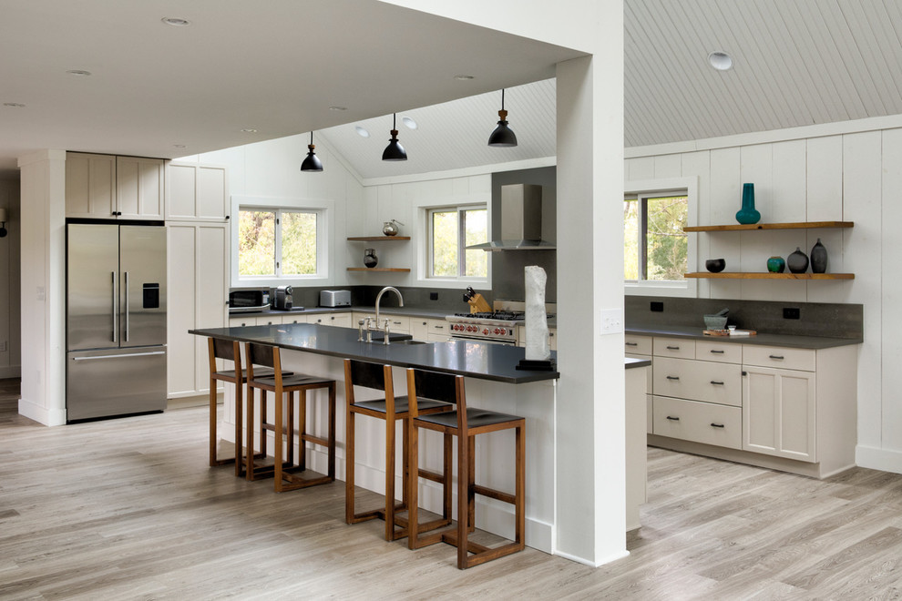 This is an example of a large country l-shaped open plan kitchen in New York with a farmhouse sink, shaker cabinets, grey cabinets, quartz benchtops, grey splashback, stone slab splashback, stainless steel appliances, light hardwood floors, with island and grey benchtop.