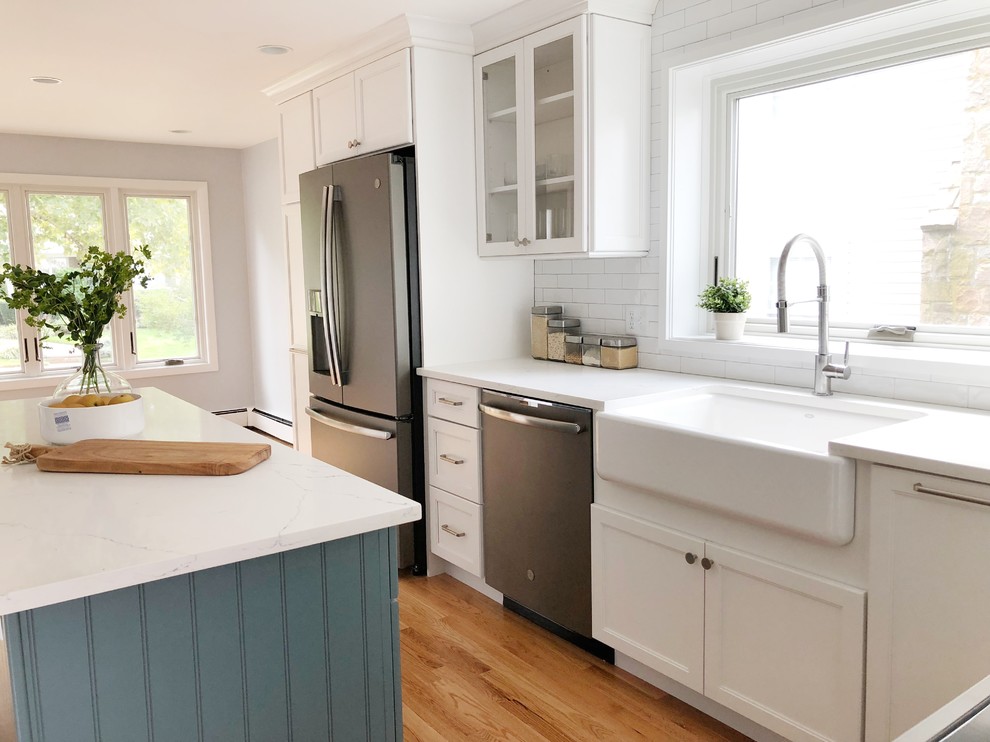 Kitchen - mid-sized transitional l-shaped light wood floor kitchen idea in Providence with a farmhouse sink, shaker cabinets, white cabinets, quartz countertops, white backsplash, ceramic backsplash, stainless steel appliances, an island and white countertops
