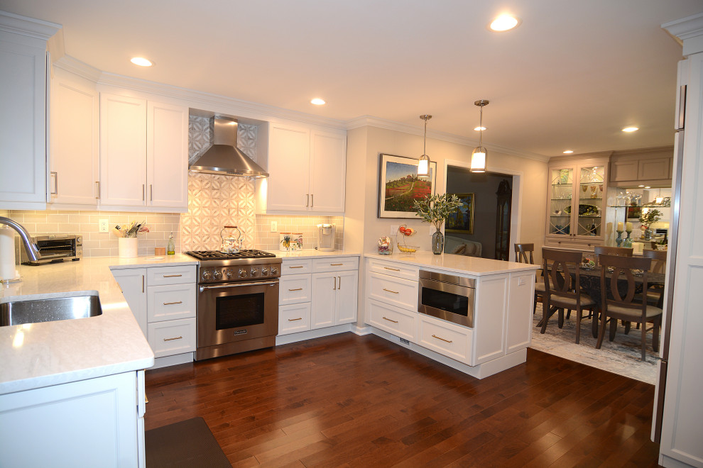 Photo of a large traditional u-shaped eat-in kitchen in New York with recessed-panel cabinets, white cabinets, quartz benchtops, beige splashback, stainless steel appliances, dark hardwood floors, a peninsula, brown floor and white benchtop.