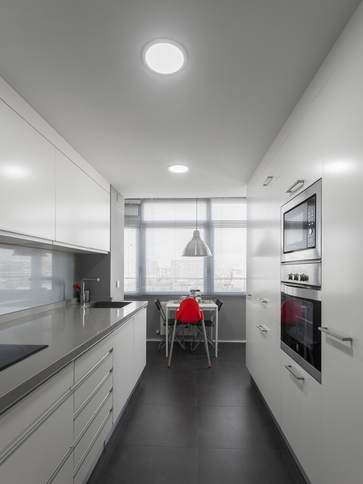 Contemporary galley separate kitchen in Valencia with a single-bowl sink, flat-panel cabinets, white cabinets, grey splashback, glass sheet splashback, black appliances, no island, black floor and grey benchtop.