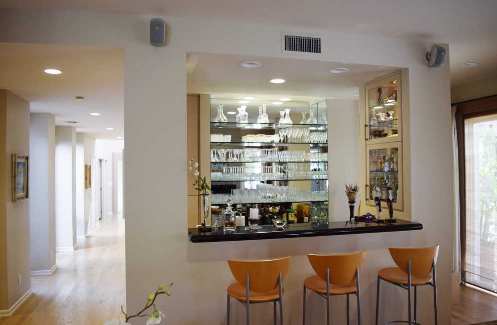 Photo of a mid-sized contemporary galley seated home bar in Los Angeles with glass-front cabinets, granite benchtops, an undermount sink, beige cabinets and light hardwood floors.