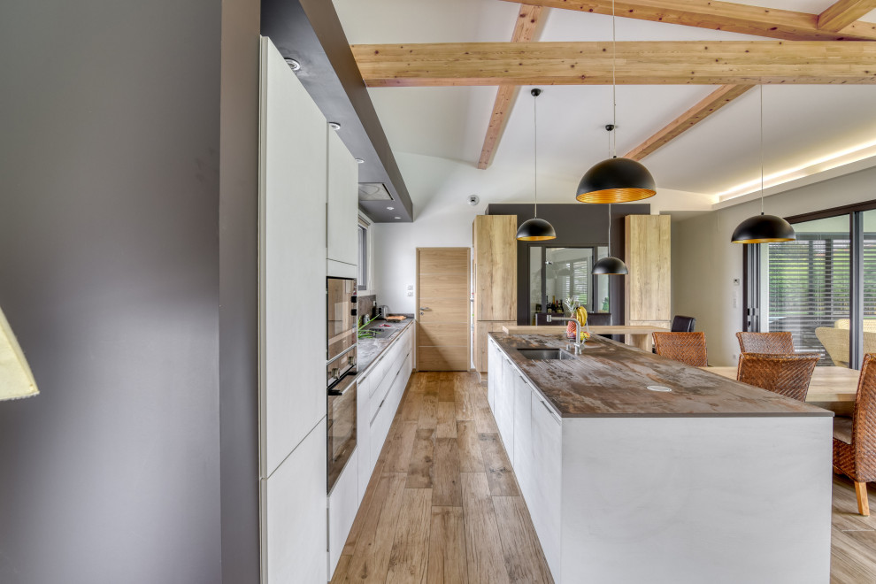 This is an example of a large industrial u-shaped open plan kitchen in Toulouse with an undermount sink, beaded inset cabinets, white cabinets, grey splashback, black appliances, light hardwood floors, with island, brown floor and grey benchtop.
