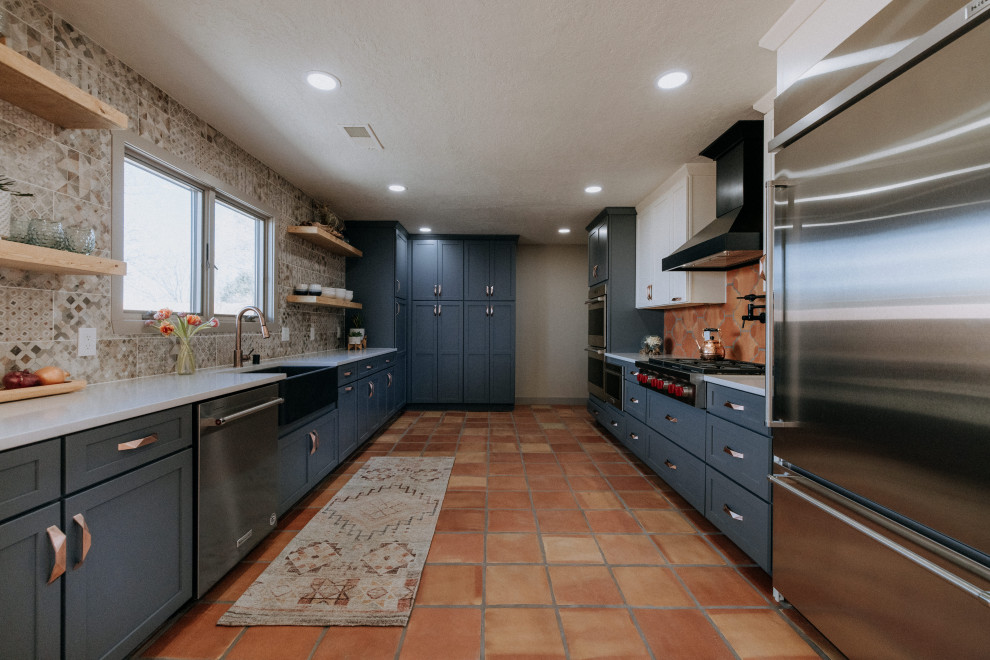Photo of a large u-shaped eat-in kitchen in Albuquerque with a farmhouse sink, shaker cabinets, blue cabinets, multi-coloured splashback, stainless steel appliances, terra-cotta floors, no island, orange floor and white benchtop.