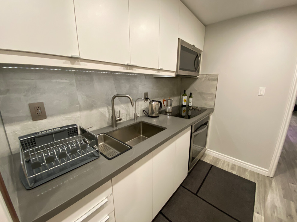 Small modern galley kitchen in Toronto with a single-bowl sink, flat-panel cabinets, white cabinets, quartz benchtops, grey splashback, porcelain splashback, stainless steel appliances, laminate floors, grey floor and grey benchtop.