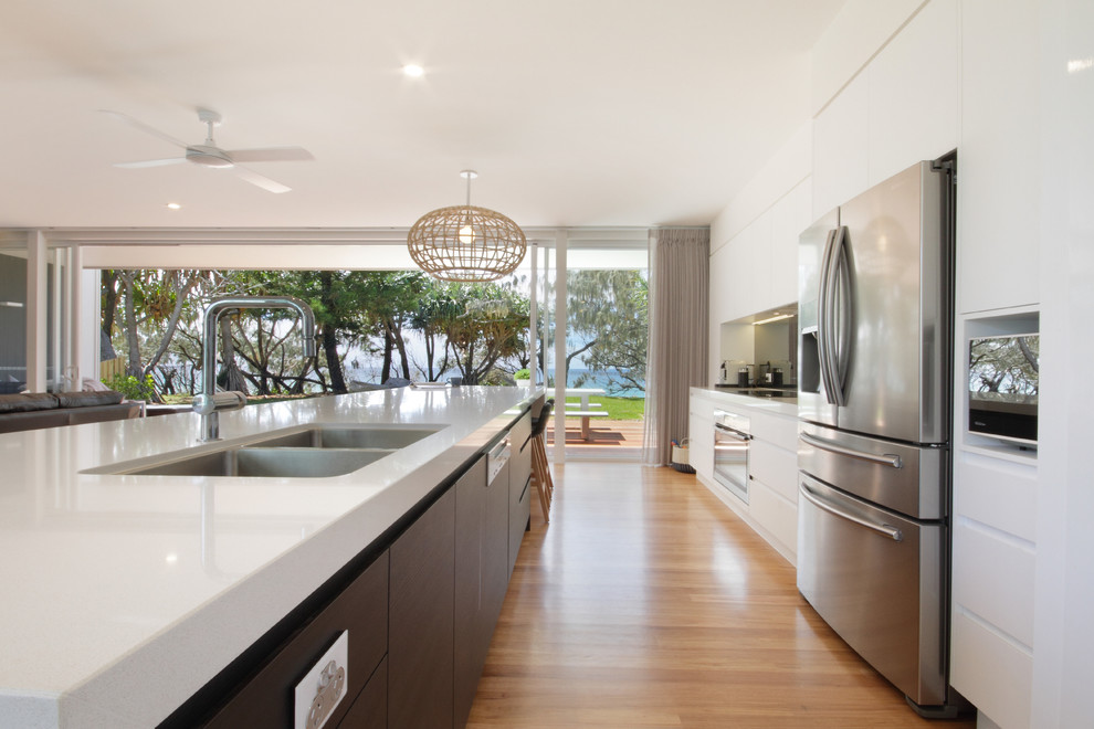This is an example of a beach style kitchen in Sunshine Coast with an undermount sink, quartz benchtops, mirror splashback, stainless steel appliances, light hardwood floors and with island.