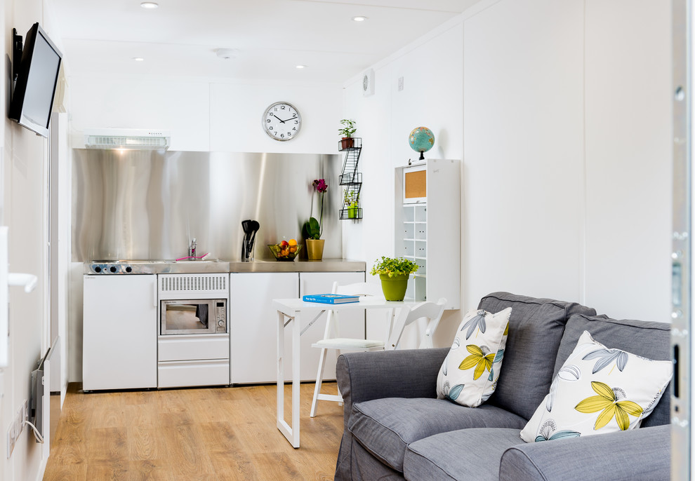 Small contemporary single-wall open plan kitchen in Manchester with an integrated sink, flat-panel cabinets, white cabinets, stainless steel benchtops, grey splashback, white appliances, medium hardwood floors, no island, brown floor and grey benchtop.