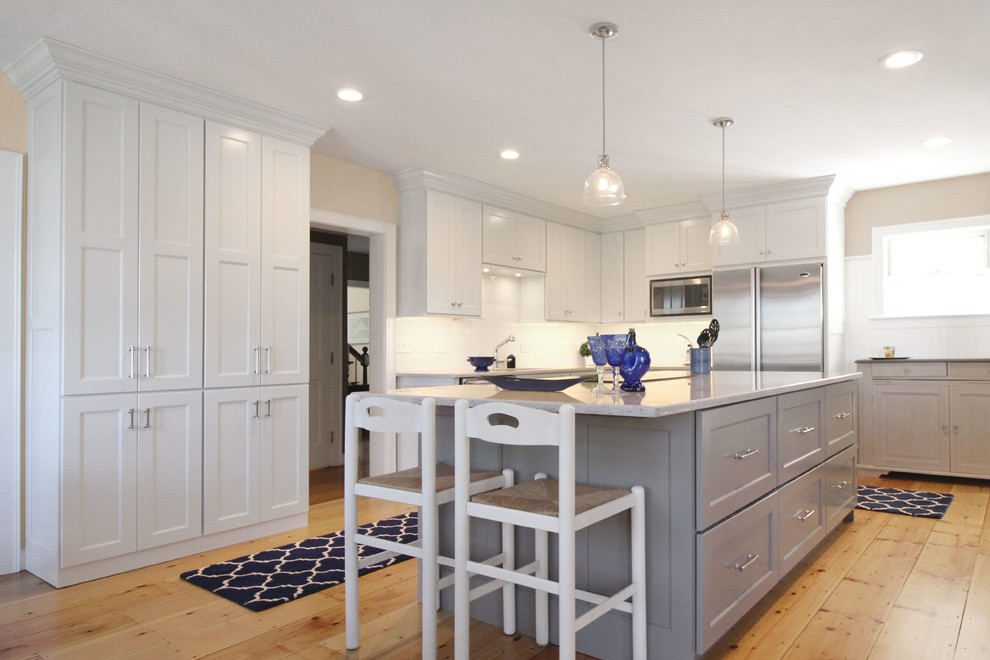 Large transitional l-shaped open plan kitchen in Boston with an undermount sink, shaker cabinets, white cabinets, quartz benchtops, white splashback, subway tile splashback, stainless steel appliances, light hardwood floors, with island and brown floor.
