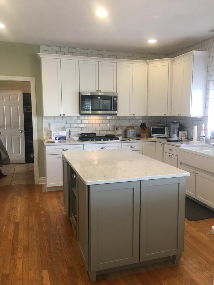 Large transitional l-shaped medium tone wood floor and brown floor open concept kitchen photo in Omaha with a farmhouse sink, shaker cabinets, white cabinets, quartz countertops, white backsplash, ceramic backsplash, stainless steel appliances, two islands and white countertops