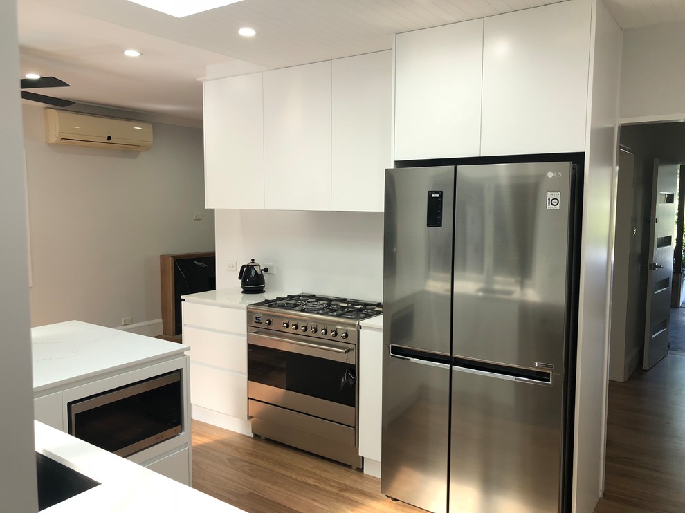 This is an example of a mid-sized contemporary u-shaped open plan kitchen in Sydney with white cabinets, stainless steel appliances, light hardwood floors, multiple islands, brown floor and white benchtop.