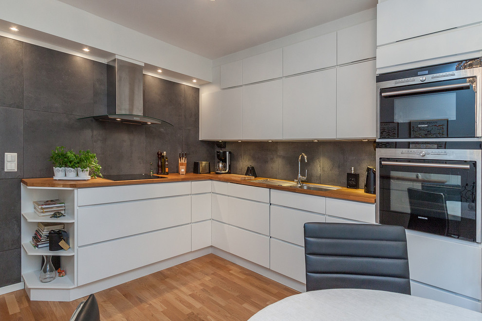 Design ideas for a contemporary l-shaped eat-in kitchen in Stockholm with a double-bowl sink, flat-panel cabinets, white cabinets, wood benchtops, black splashback, stainless steel appliances, light hardwood floors and no island.