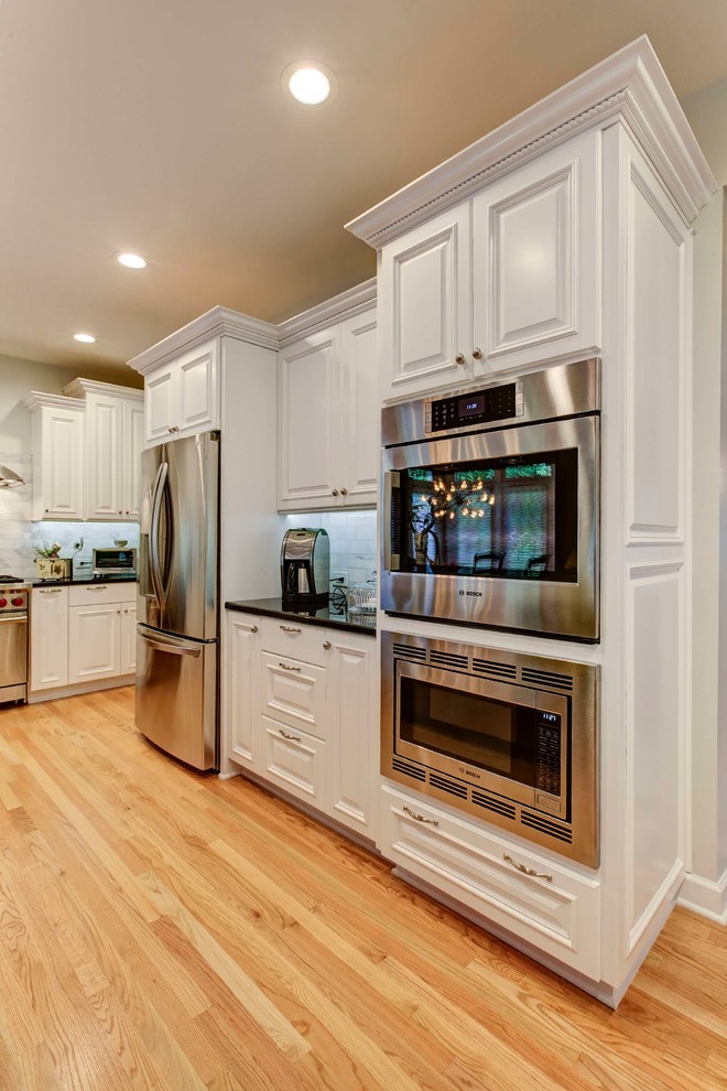 Photo of a mid-sized transitional galley eat-in kitchen in Raleigh with a double-bowl sink, raised-panel cabinets, white cabinets, granite benchtops, white splashback, stone tile splashback, stainless steel appliances, light hardwood floors and with island.