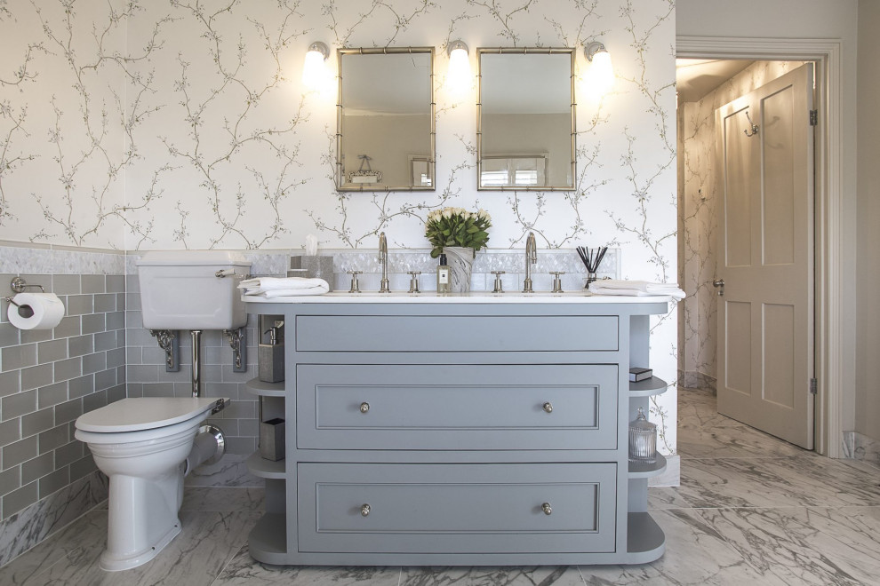 This is an example of a transitional master bathroom in London with recessed-panel cabinets, grey cabinets, a two-piece toilet, gray tile, subway tile, multi-coloured walls, marble floors, an undermount sink, grey floor, white benchtops, a double vanity, a built-in vanity and wallpaper.