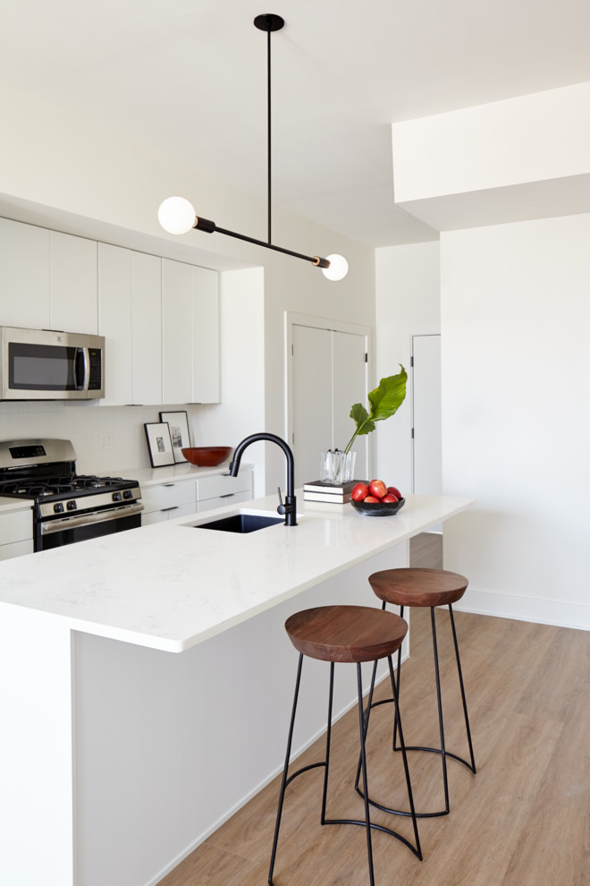 Small contemporary galley open plan kitchen in Philadelphia with an undermount sink, flat-panel cabinets, white cabinets, quartz benchtops, white splashback, ceramic splashback, stainless steel appliances, light hardwood floors, with island and beige floor.