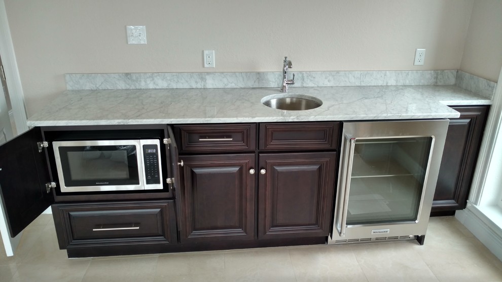 Photo of a small beach style galley wet bar in Miami with an undermount sink, raised-panel cabinets, dark wood cabinets, marble benchtops, white splashback and travertine floors.