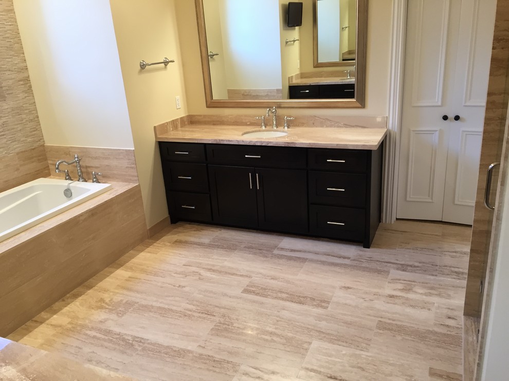 This is an example of a large modern master bathroom in Dallas with an undermount sink, shaker cabinets, dark wood cabinets, marble benchtops, a drop-in tub, an alcove shower, a two-piece toilet, beige tile, stone tile, beige walls and travertine floors.