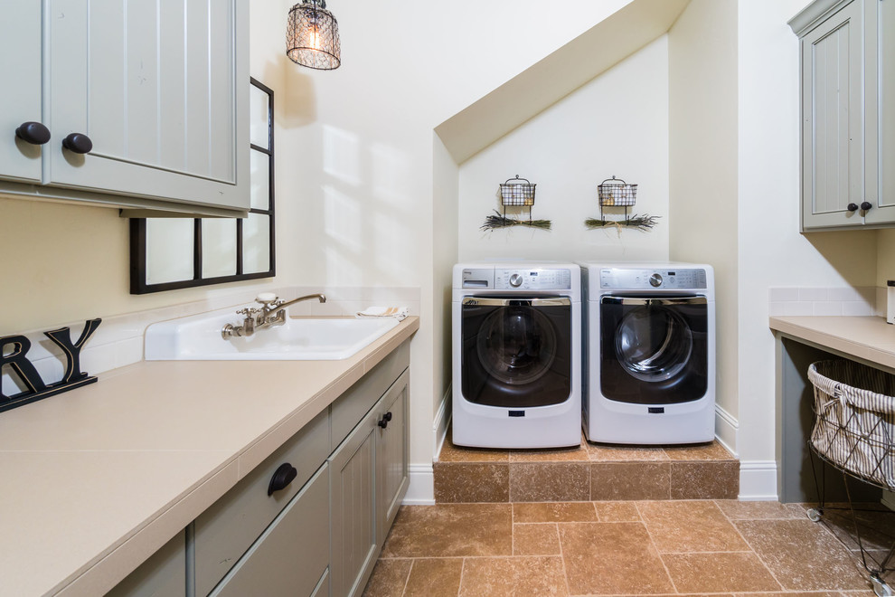 Design ideas for a mid-sized mediterranean galley laundry room in Other with a drop-in sink, grey cabinets, laminate benchtops, beige walls, limestone floors, a side-by-side washer and dryer, brown floor and recessed-panel cabinets.