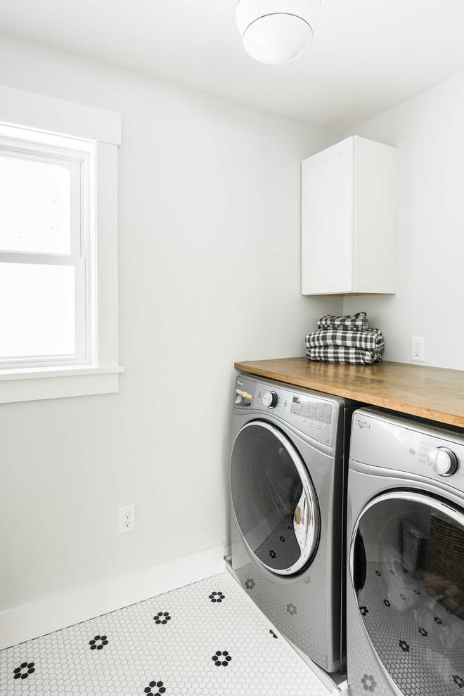 Mid-sized transitional dedicated laundry room in Portland Maine with shaker cabinets, white cabinets, wood benchtops, porcelain floors, a side-by-side washer and dryer, white floor and grey walls.