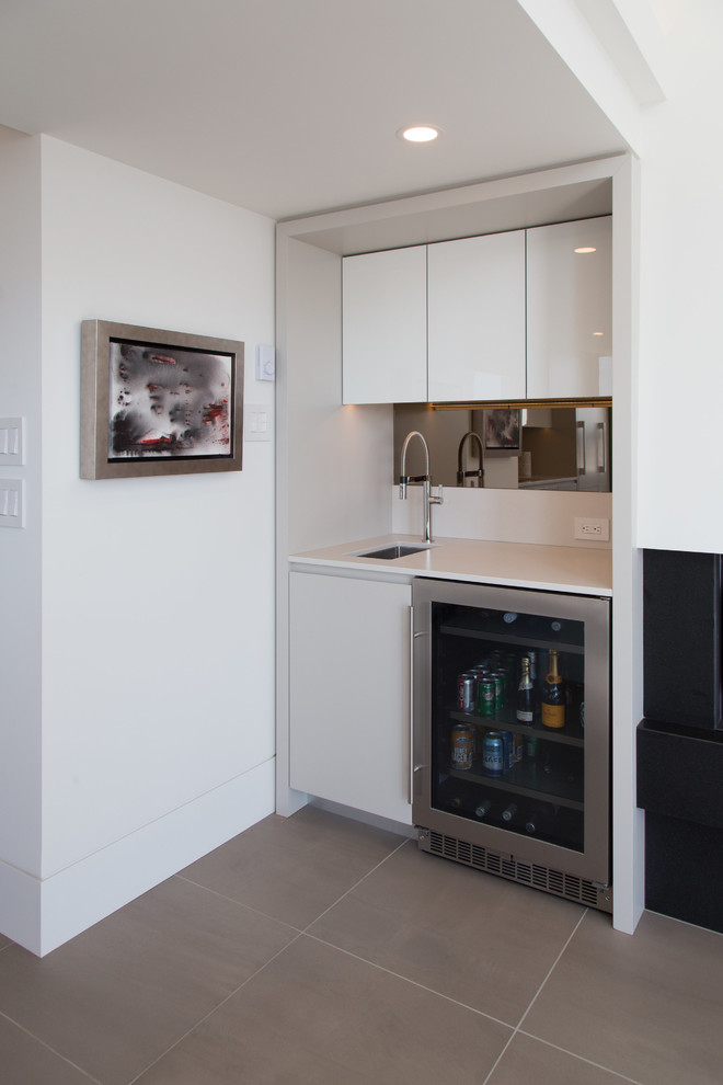 Small contemporary single-wall wet bar in Vancouver with an undermount sink, flat-panel cabinets, white cabinets, solid surface benchtops, mirror splashback, porcelain floors, beige floor and white benchtop.