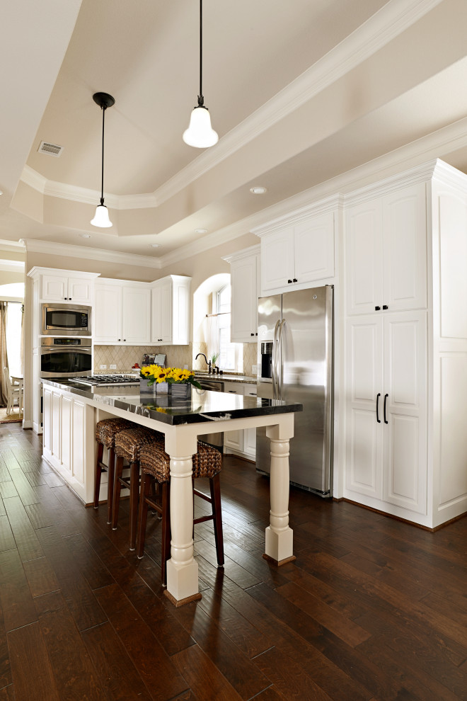 Design ideas for a mid-sized traditional u-shaped eat-in kitchen in Dallas with a double-bowl sink, white cabinets, granite benchtops, beige splashback, porcelain splashback, stainless steel appliances, medium hardwood floors, with island, brown floor and black benchtop.