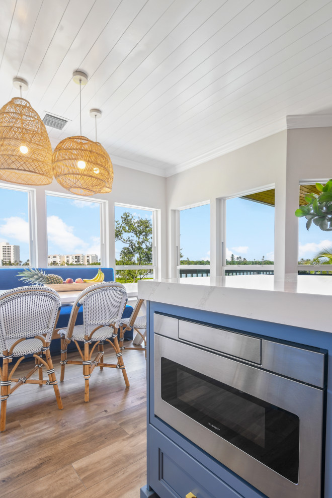 This is an example of a mid-sized beach style l-shaped open plan kitchen in Other with a farmhouse sink, shaker cabinets, blue cabinets, quartz benchtops, white splashback, porcelain splashback, black appliances, porcelain floors, with island, brown floor, white benchtop and timber.