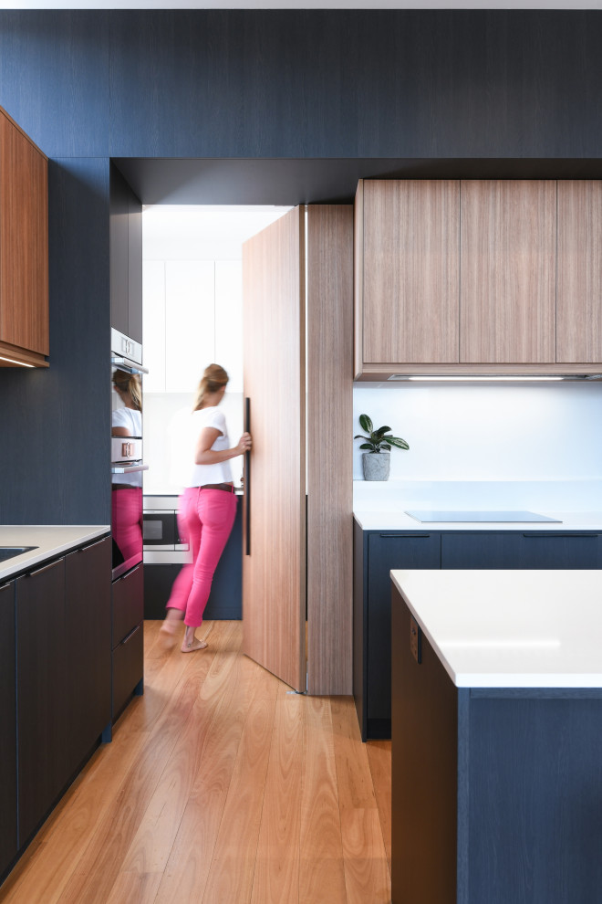 This is an example of a large modern l-shaped open plan kitchen in Sydney with a drop-in sink, flat-panel cabinets, black cabinets, quartz benchtops, white splashback, stone slab splashback, black appliances, medium hardwood floors, with island, brown floor and white benchtop.