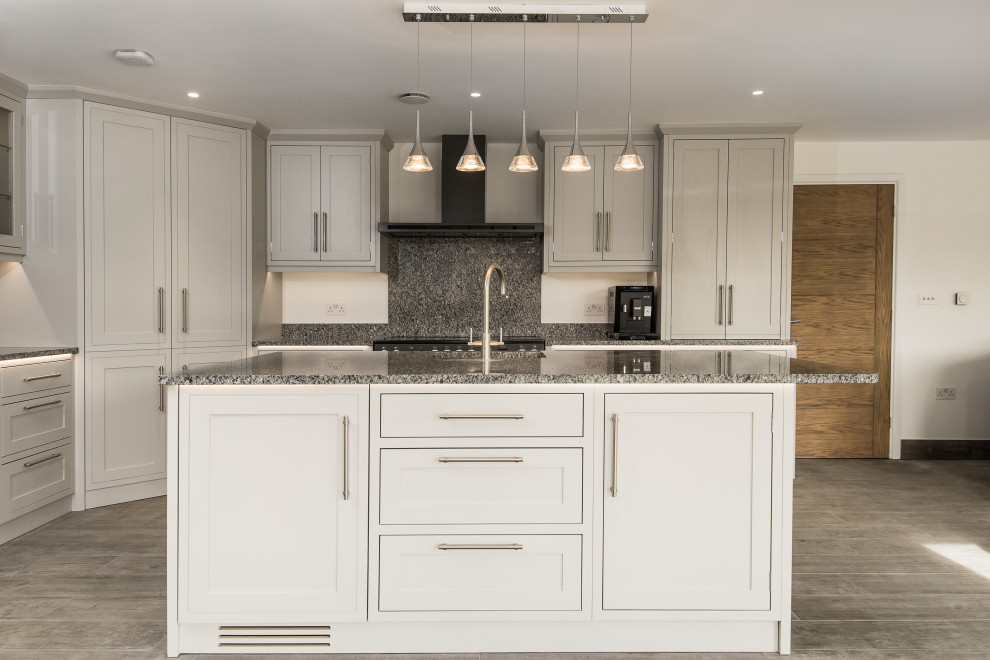 This is an example of a traditional l-shaped eat-in kitchen in Oxfordshire with a drop-in sink, shaker cabinets, grey cabinets, granite benchtops, grey splashback, granite splashback, black appliances, with island, grey floor and grey benchtop.