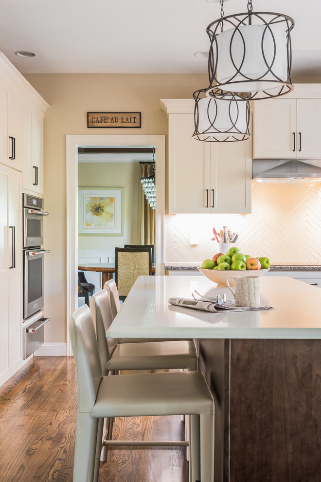 Example of a transitional medium tone wood floor and brown floor kitchen design in Boston with shaker cabinets, white cabinets, quartzite countertops, beige backsplash, glass tile backsplash, stainless steel appliances and an island