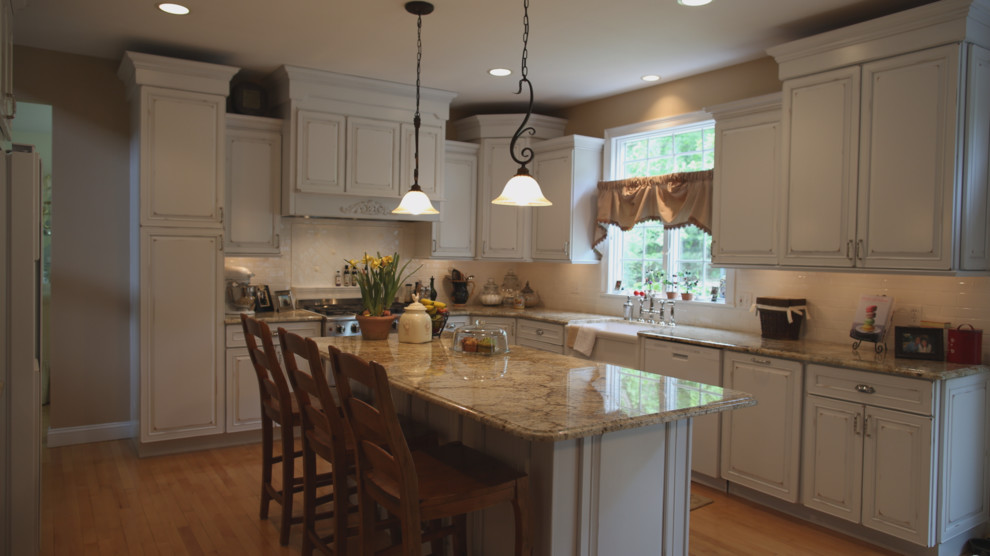 This is an example of a traditional kitchen in Providence with a farmhouse sink, raised-panel cabinets, distressed cabinets, granite benchtops, white splashback, ceramic splashback, white appliances, medium hardwood floors and with island.