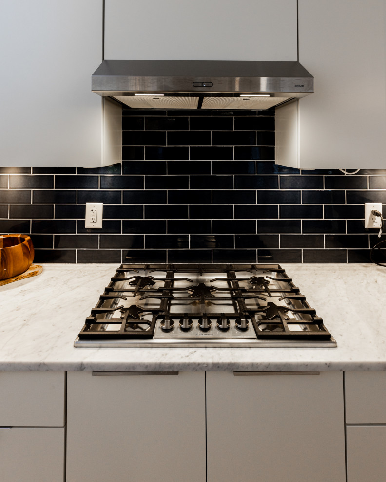 This is an example of a large scandi galley kitchen/diner in Atlanta with a submerged sink, flat-panel cabinets, beige cabinets, marble worktops, black splashback, metro tiled splashback, stainless steel appliances, light hardwood flooring, an island, brown floors and white worktops.