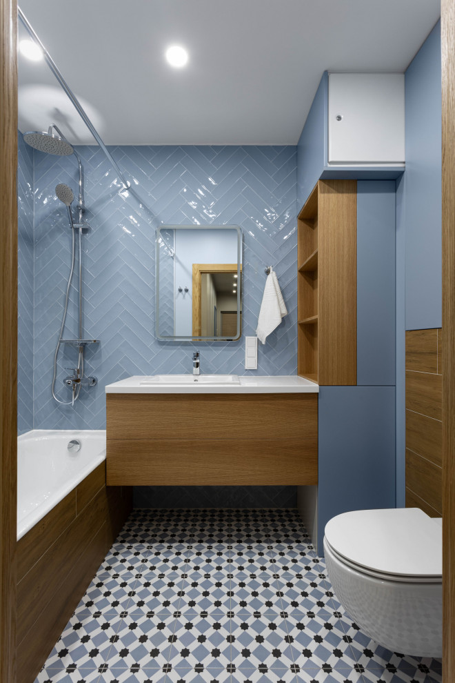 This is an example of a medium sized scandinavian ensuite bathroom in Saint Petersburg with flat-panel cabinets, medium wood cabinets, a built-in bath, a wall mounted toilet, blue tiles, ceramic tiles, blue walls, ceramic flooring, a submerged sink, solid surface worktops, grey floors, white worktops, a single sink and a floating vanity unit.
