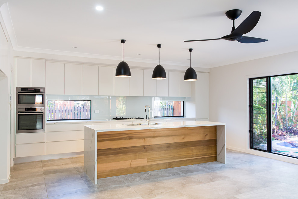 Photo of a contemporary galley open plan kitchen in Townsville with a double-bowl sink, flat-panel cabinets, white cabinets, marble benchtops, white splashback, glass sheet splashback, stainless steel appliances, ceramic floors, with island and grey floor.